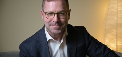 Man in a navy check blazer and white shirt sits on a sofa, smiling at the camera with a warm lamp in the background.