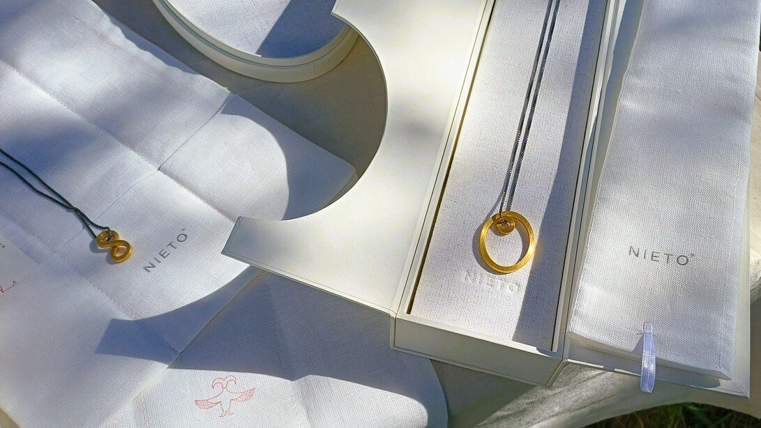 Gold circular pendant necklace displayed on white fabric with Nieto branding on boxes.