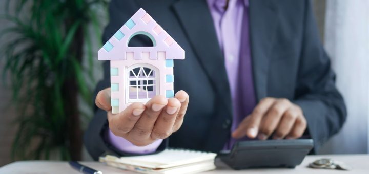 Person in a suit holds a pastel toy house while using a calculator at a desk with papers and a pen nearby.