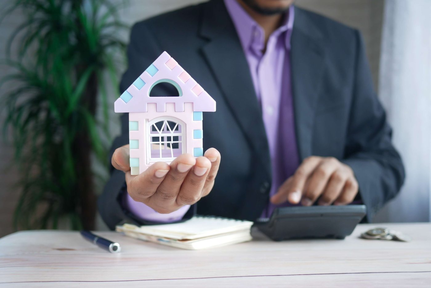 Person in a suit holds a pastel toy house while using a calculator at a desk with papers and a pen nearby.
