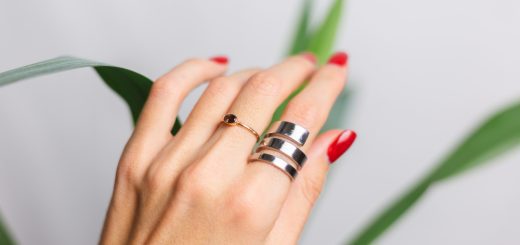 Close-up of a hand with red nails wearing two rings—a copper ring with a small gemstone and a stacked silver ring—against a blurred plant backdrop.