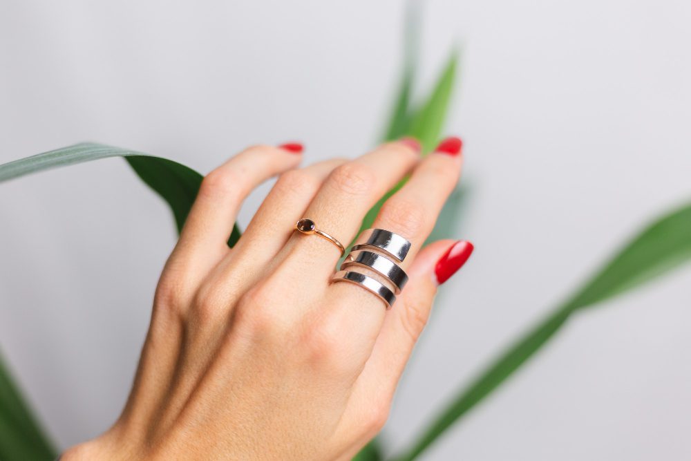 Close-up of a hand with red nails wearing two rings—a copper ring with a small gemstone and a stacked silver ring—against a blurred plant backdrop.