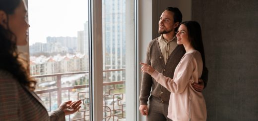 A woman on the left explains features to a couple standing by a large window in a high-rise apartment, with city buildings outside.