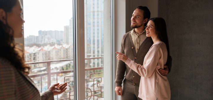 A woman on the left explains features to a couple standing by a large window in a high-rise apartment, with city buildings outside.