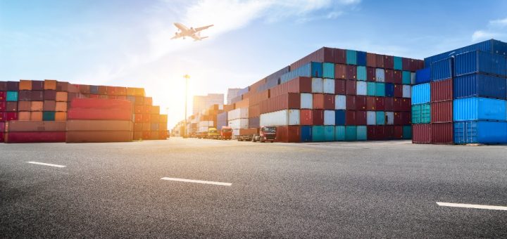 Port yard with colorful shipping containers, trucks, and an airplane taking off in a clear blue sky.