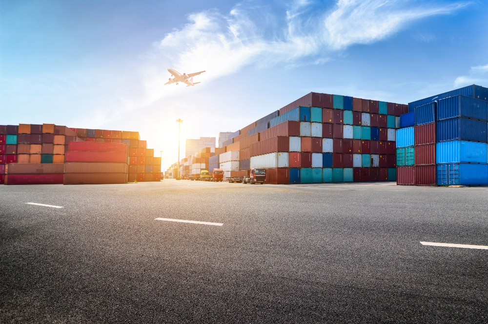 772686 - VSI Consulting - EconomiaeInversion.com Port yard with colorful shipping containers, trucks, and an airplane taking off in a clear blue sky.
