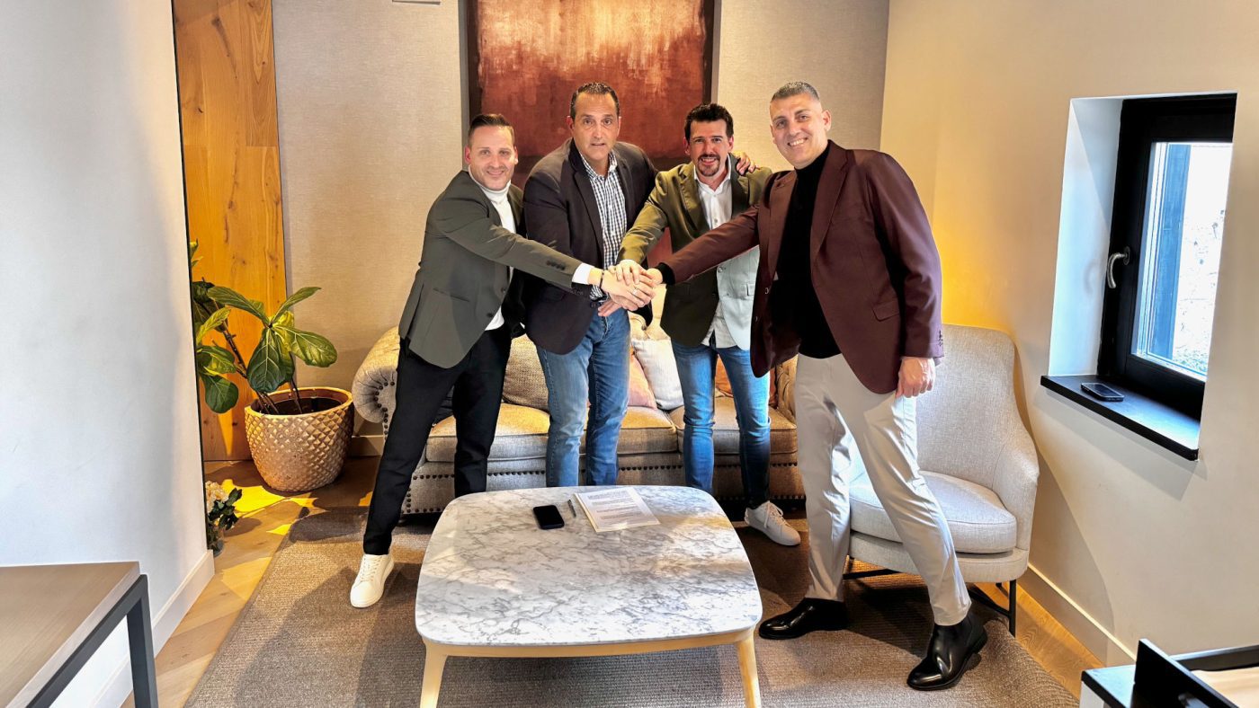 Four men in blazers shake hands in a modern living room around a marble coffee table.