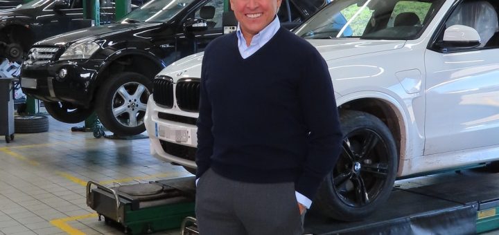 Smiling man in a navy sweater stands in an automotive repair shop with a white SUV on a lift behind him in the foreground.