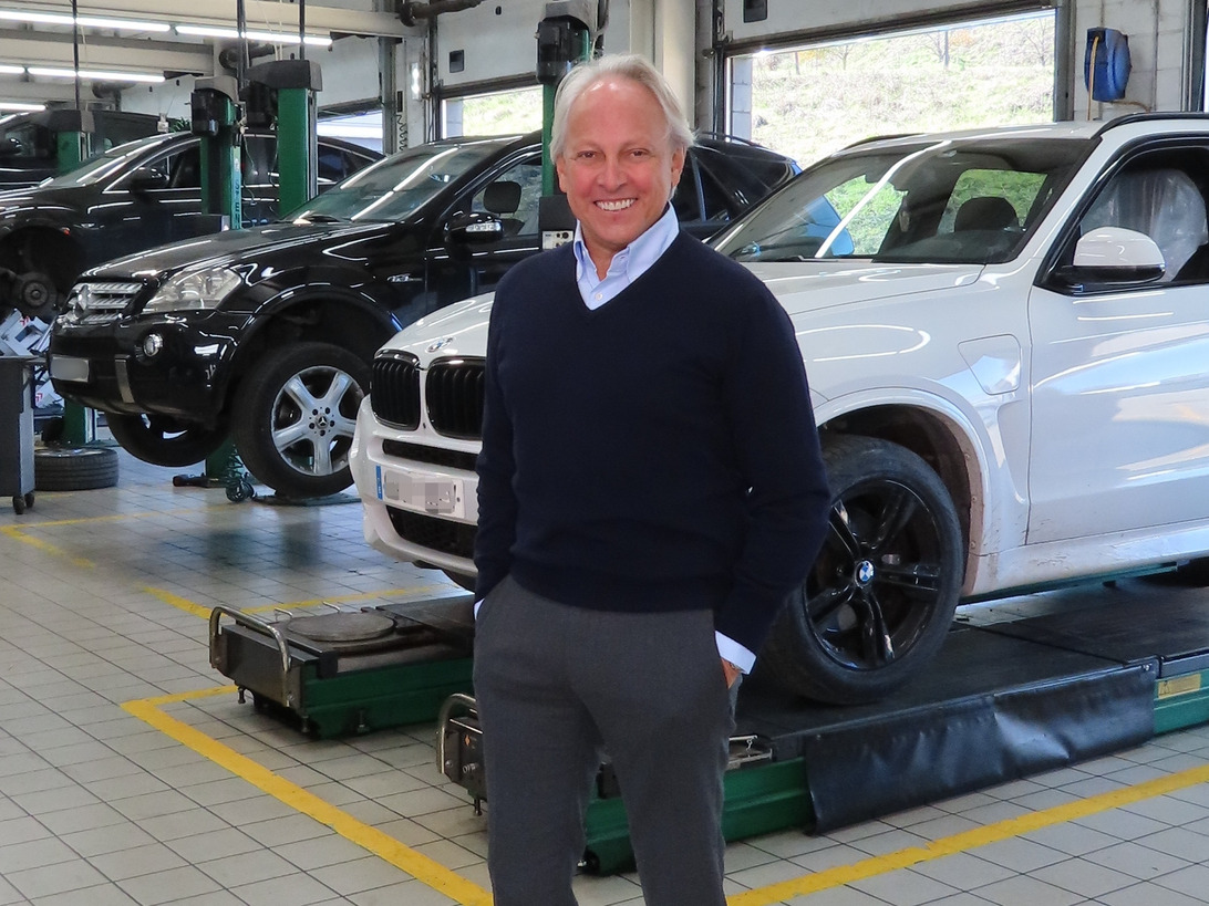 Smiling man in a navy sweater stands in an automotive repair shop with a white SUV on a lift behind him in the foreground.