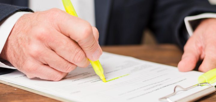 Person in a suit signs a document with a bright yellow highlighter on a clipboard (close-up).