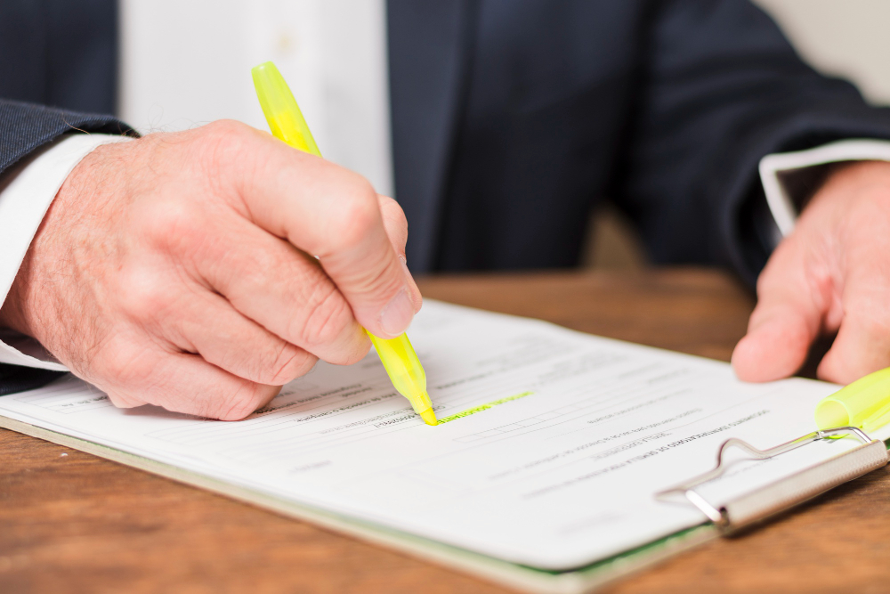 Person in a suit signs a document with a bright yellow highlighter on a clipboard (close-up).