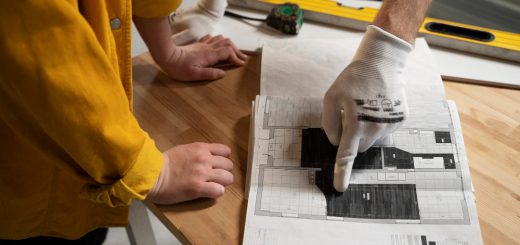 Worker wearing a glove points to a detailed architectural blueprint laid on a wooden table, with another hand nearby supporting the paper.
