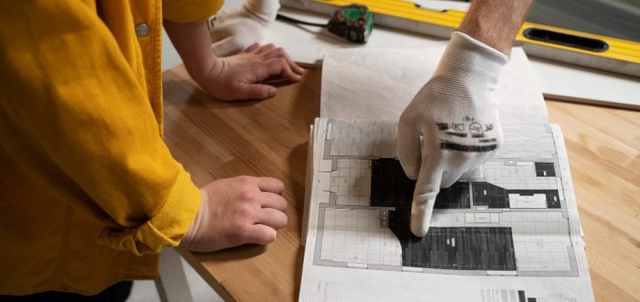 Worker wearing a glove points to a detailed architectural blueprint laid on a wooden table, with another hand nearby supporting the paper.