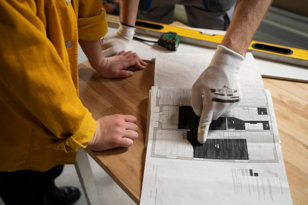 Worker wearing a glove points to a detailed architectural blueprint laid on a wooden table, with another hand nearby supporting the paper.