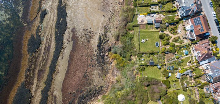 Aerial view of a coastal neighborhood: rocky shoreline on the left, houses and green gardens on the right along a street with parked cars.