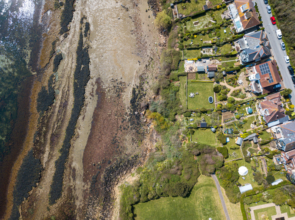 Aerial view of a coastal neighborhood: rocky shoreline on the left, houses and green gardens on the right along a street with parked cars.