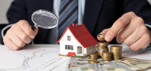 Businessperson in a suit examines a miniature house with a magnifying glass, coins, and charts on a desk for real estate investment analysis