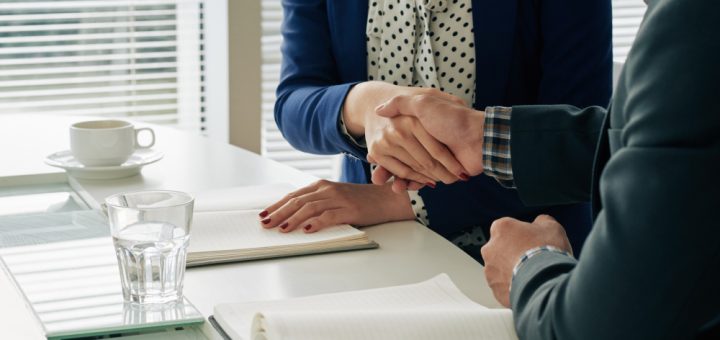 Two professionals shake hands over a white table with notebooks and a glass of water, signaling an agreement or closing a deal.
