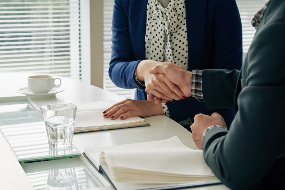 773732 - Briones Abogados - EconomiaeInversion.com Two professionals shake hands over a white table with notebooks and a glass of water, signaling an agreement or closing a deal.