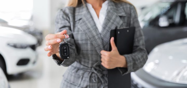 Woman in a checkered blazer handing over car key fob in a showroom, clipboard in hand