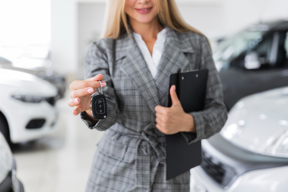 774684 - Vasa Taldea - EconomiaeInversion.com Woman in a checkered blazer handing over car key fob in a showroom, clipboard in hand