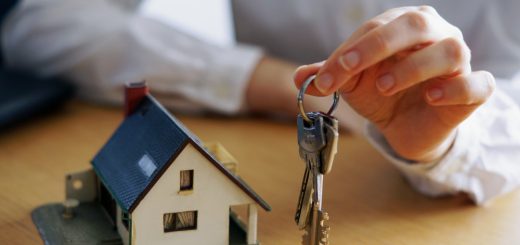 Hand holding a key ring with several keys beside a small model house on a wooden table, symbolizing home ownership or real estate.