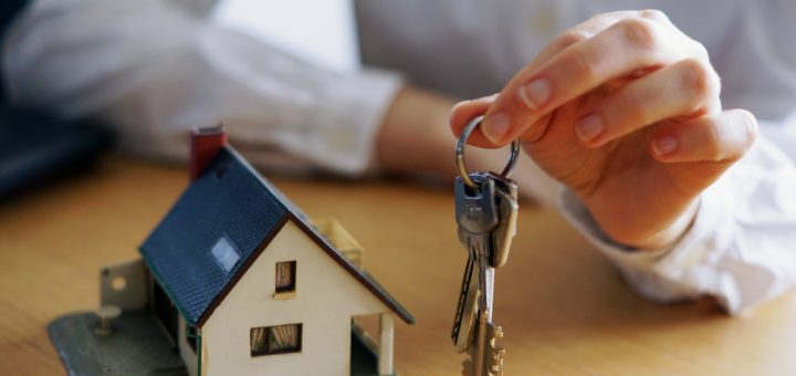 Hand holding a key ring with several keys beside a small model house on a wooden table, symbolizing home ownership or real estate.