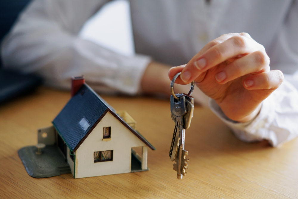775144 - Gestibensa - EconomiaeInversion.com Hand holding a key ring with several keys beside a small model house on a wooden table, symbolizing home ownership or real estate.