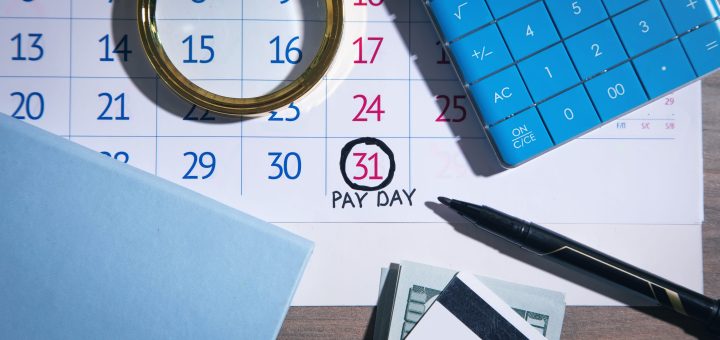 Calendar page with the 31st circled and 'PAY DAY' written beneath, surrounded by a magnifying glass, a blue calculator, a pen, and cash on a desk.