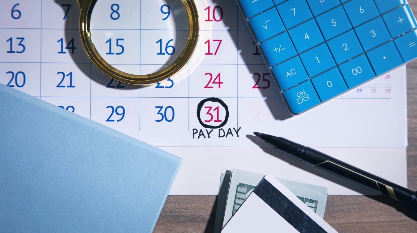Pay day of the month Marker calculator notepad on calendar - EconomiaeInversion.com Calendar page with the 31st circled and 'PAY DAY' written beneath, surrounded by a magnifying glass, a blue calculator, a pen, and cash on a desk.