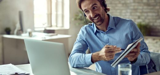 Man with a headset at a desk, smiling while pointing at a tablet beside an open laptop in a bright office setting