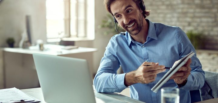Man with a headset at a desk, smiling while pointing at a tablet beside an open laptop in a bright office setting