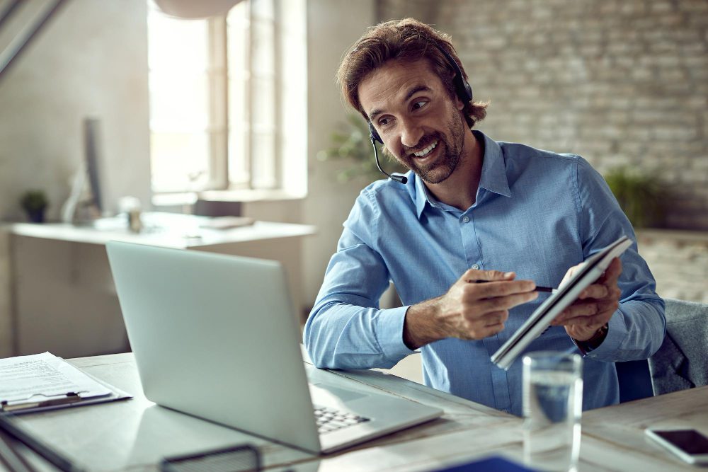 Man with a headset at a desk, smiling while pointing at a tablet beside an open laptop in a bright office setting