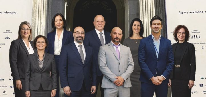 Group of eight professionals in business attire posing for a photo on stone steps in front of a historic building, with white banners displaying logos on both sides.