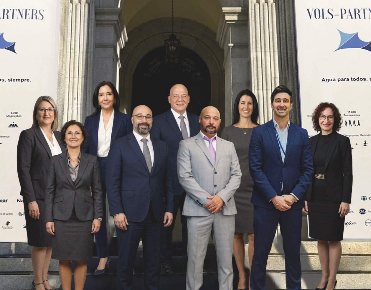 Group of eight professionals in business attire posing for a photo on stone steps in front of a historic building, with white banners displaying logos on both sides.
