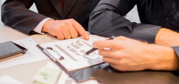 Two professionals review a document on a clipboard, one pointing with a pen at a chart.