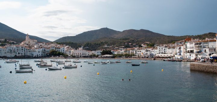 Harbor in a Mediterranean town with white buildings, boats on calm water, and hills in the background