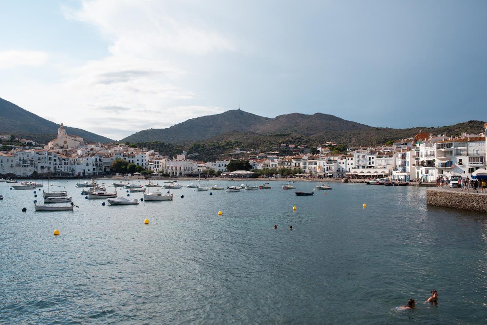 776266 - Soms Granados - EconomiaeInversion.com Harbor in a Mediterranean town with white buildings, boats on calm water, and hills in the background
