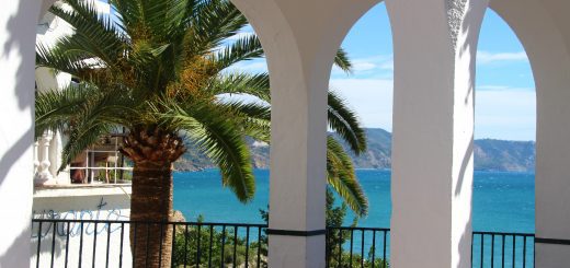 White arched terrace framing a palm tree and blue sea beyond, with a black railing in the foreground and distant hills under a clear sky.
