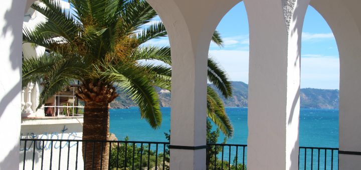 White arched terrace framing a palm tree and blue sea beyond, with a black railing in the foreground and distant hills under a clear sky.