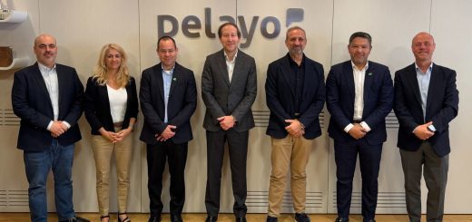 Seven professionals in business attire standing in a row for a group photo in a modern office, with a company logo on the wall behind them.