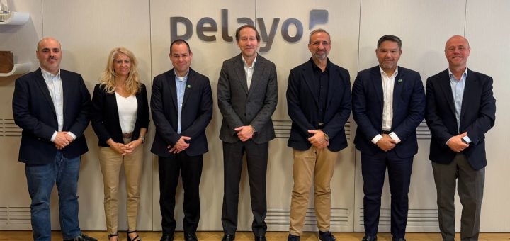 Seven professionals in business attire standing in a row for a group photo in a modern office, with a company logo on the wall behind them.