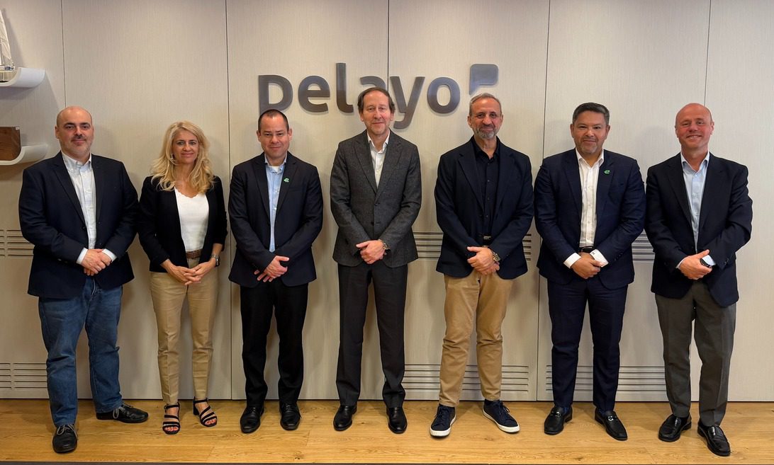 Seven professionals in business attire standing in a row for a group photo in a modern office, with a company logo on the wall behind them.