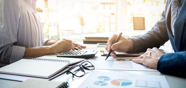 Two professionals in a bright office review charts and graphs across a desk, using a calculator and taking notes.