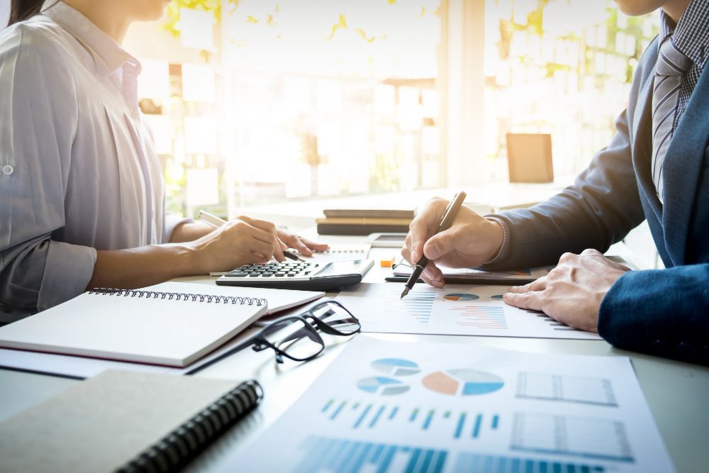 Two professionals in a bright office review charts and graphs across a desk, using a calculator and taking notes.