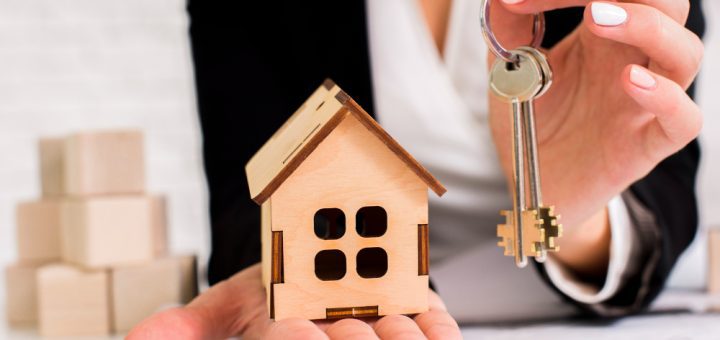 A person holding a small wooden house model in one hand and a set of keys in the other, symbolizing home ownership or real estate.