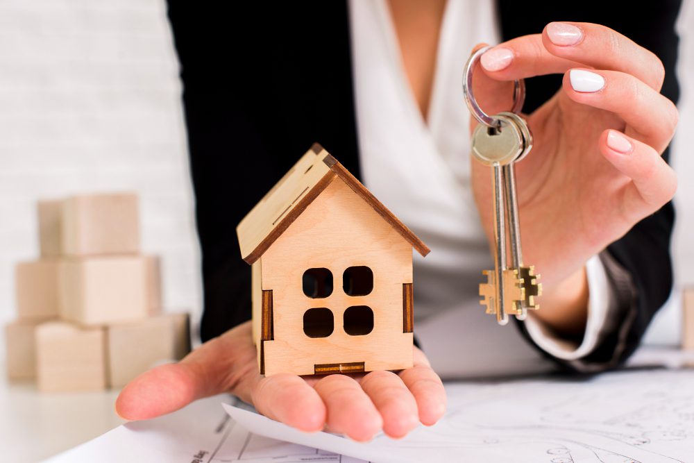 A person holding a small wooden house model in one hand and a set of keys in the other, symbolizing home ownership or real estate.