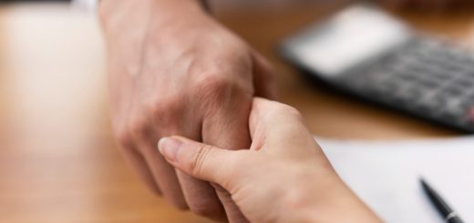 Two hands in a handshake over a wooden desk, signaling a business agreement or greeting.